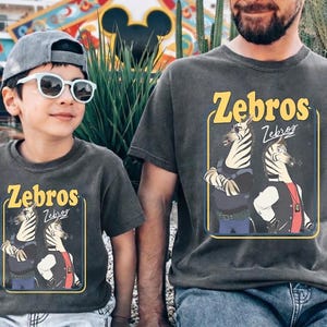 May include: Two charcoal gray t-shirts with the word "Zebros" in yellow, featuring a cartoon zebra design. One shirt is worn by a child wearing sunglasses and a cap. The other is worn by an adult.