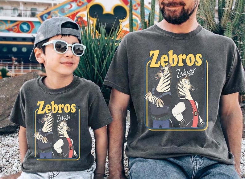 May include: Two charcoal gray t-shirts with the word "Zebros" in yellow, featuring a cartoon zebra design. One shirt is worn by a child wearing sunglasses and a cap. The other is worn by an adult.