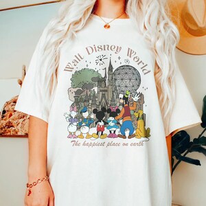 May include: Cream-colored t-shirt featuring a colorful graphic of Disney characters and landmarks, including Mickey and Minnie Mouse, Goofy, and Cinderella's castle. The text "Walt Disney World" and "The happiest place on earth" are printed above and below the illustration.