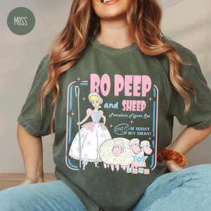 以下が含まれることがあります： モスグリーンのTシャツ。トイ・ストーリーのボー・ピープと羊のグラフィック。デザインには、ピンクと青で「RO PEEP and SHEEP Porcelain Figure Set」と「ONLY AT SELRCT TOY STORY！」の文字が含まれています。