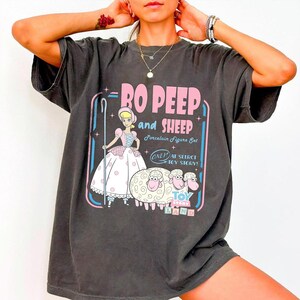 以下が含まれることがあります： トイ・ストーリーのボー・ピープと羊のグラフィックが特徴の濃いグレーのTシャツ。「RO PEEP and SHEEP Porcelain Figure Set」の文字がピンクと白でデザインされ、キャラクターの漫画イラストが描かれています。ヴィンテージ感のあるシャツです。