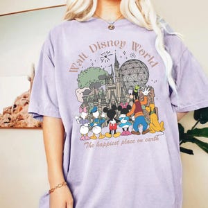 May include: A light purple t-shirt featuring a graphic of Disney World characters and landmarks. The design includes Mickey and Minnie Mouse, Donald and Daisy Duck, Goofy, and Pluto. Text reads "Walt Disney World" and "The happiest place on earth."