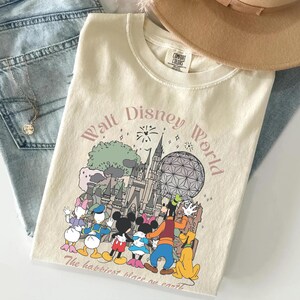 May include: Cream-colored t-shirt featuring a graphic of Disney characters and landmarks, including Cinderella's castle and Spaceship Earth. The text "Walt Disney World" and "The happiest place on earth" are printed on the shirt. A tan hat and blue jeans are in the background.