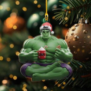 May include: A Christmas ornament featuring the Hulk in a Santa hat with the name "Robert". The green figure holds a red mug with "2025" and a gingerbread man. The ornament is suspended by a gold string.