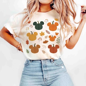 May include: Cream-colored t-shirt featuring a pattern of pumpkins with mouse ear-like shapes, along with fall leaves and sprigs. The pumpkins are in shades of orange, brown, and green. The shirt is a casual, short-sleeved style.