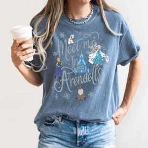 Puede incluir: Una camiseta azul con el texto "Meet me in Arendelle" en una fuente caprichosa. La camiseta presenta ilustraciones de personajes y elementos de la pel&iacute;cula Frozen, incluyendo Elsa, Anna y Olaf.