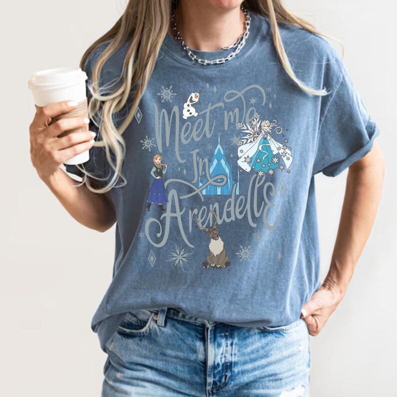 Puede incluir: Una camiseta azul con el texto "Meet me in Arendelle" en una fuente caprichosa. La camiseta presenta ilustraciones de personajes y elementos de la pel&iacute;cula Frozen, incluyendo Elsa, Anna y Olaf.