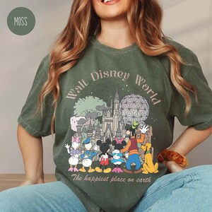 May include: Moss green t-shirt featuring a graphic of Walt Disney World characters and landmarks. The design includes Mickey and Minnie Mouse, Donald and Daisy Duck, Goofy, Pluto, and Cinderella's castle. Text reads "Walt Disney World" and "The happiest place on earth."