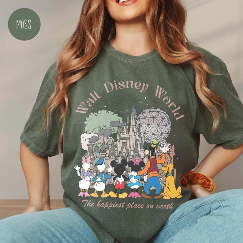 May include: Moss green t-shirt featuring a graphic of Walt Disney World characters and landmarks. The design includes Mickey and Minnie Mouse, Donald and Daisy Duck, Goofy, Pluto, and Cinderella's castle. Text reads "Walt Disney World" and "The happiest place on earth."
