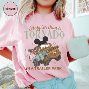 May include: Pink t-shirt with the text "Happier than a TORNADO in a Trailer Park" and a cartoon image of Mater from the Cars movie with Mickey Mouse ears. The shirt has a crew neck and short sleeves.