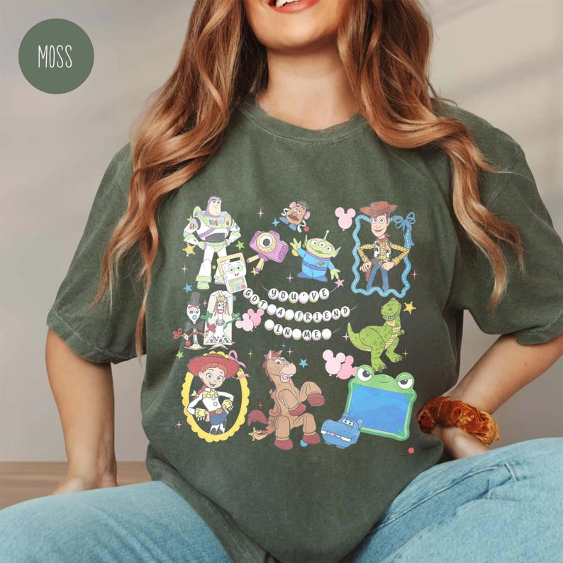 May include: A moss green short-sleeve t-shirt featuring a graphic print of various Toy Story characters including Woody, Buzz Lightyear, Jessie, and Rex. The design includes the text "YOU'VE GOT A FRIEND IN ME" arranged in a circular pattern.