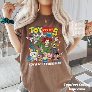 May include: Espresso-colored t-shirt featuring a graphic of Toy Story characters with the text "TOY STORY 5" and "YOU'VE GOT A FRIEND IN ME." The shirt is from Comfort Colors.