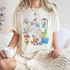 May include: A cream-colored t-shirt featuring a colorful graphic print of various animated toy characters, including a cowboy, a space ranger, a dinosaur, and a cowgirl, with the text "YOU'VE GOT A FRIEND IN ME" arranged in a circular pattern.