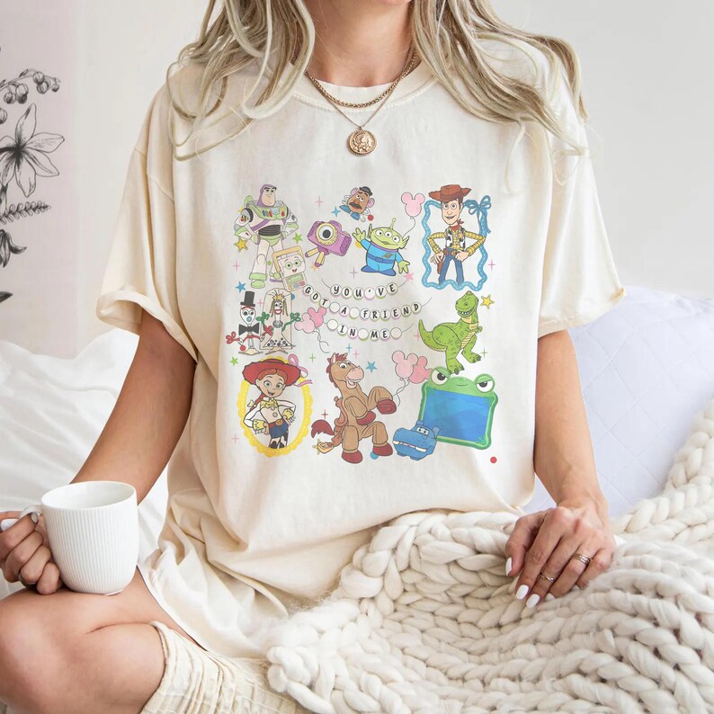 May include: A cream-colored t-shirt featuring a colorful graphic print of various animated toy characters, including a cowboy, a space ranger, a dinosaur, and a cowgirl, with the text "YOU'VE GOT A FRIEND IN ME" arranged in a circular pattern.