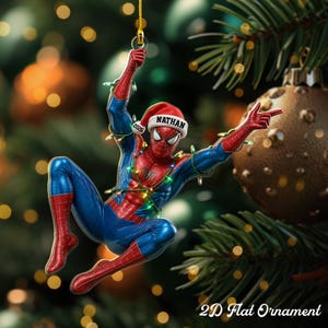 May include: A 2D flat Christmas ornament featuring a red and blue superhero character wearing a Santa hat with the name NATHAN and the year 2026 printed on it. The character is tangled in colorful string lights while hanging from a gold cord.