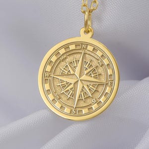 Puede incluir: Collar de oro con un colgante redondo con un diseño de rosa de los vientos. El colgante tiene las direcciones cardinales N, S, E y W grabadas alrededor del borde.