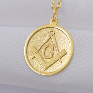 Könnte beinhalten: Goldfarbene Halskette mit rundem Anhänger. Der Anhänger hat einen strukturierten Hintergrund und zeigt einen Zirkel und ein Winkelmaß mit dem Buchstaben "G" in der Mitte. Die Kette ist aus feinem Gold gefertigt.