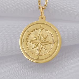 May include: A gold-coloured compass pendant necklace. The pendant features a compass rose design with directional letters. The necklace hangs on a delicate chain, set against a white fabric background. The pendant is round and has a polished finish.