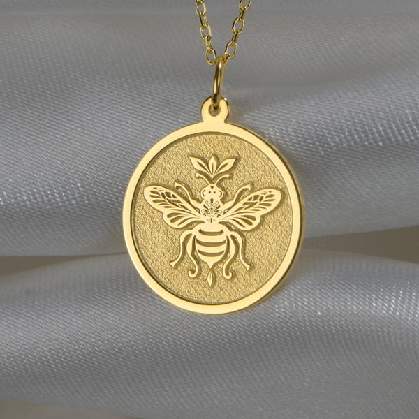 May include: A gold-toned necklace featuring a circular pendant with a detailed embossed bee design. The pendant is suspended from a delicate gold-toned chain. The bee motif is centered within the round frame, showcasing intricate wing and body details.