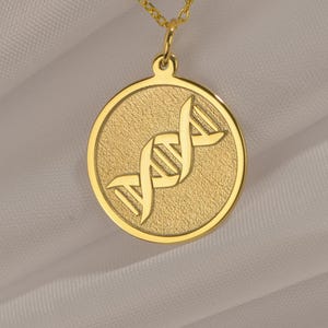 Può includere: Una collana color oro con un ciondolo circolare dalla superficie testurizzata e un design a doppia elica del DNA in rilievo al centro. Il ciondolo è sospeso a una delicata catenina color oro, creando un gioiello semplice e coerente.
