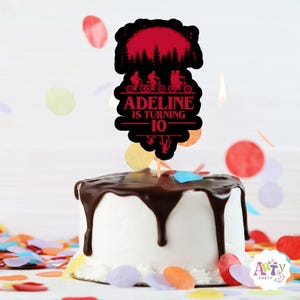 Puede incluir: Una tarta de cumpleaños con glaseado blanco y ganache de chocolate. Un adorno para tarta negro y rojo que dice "Adeline cumple 10" con una silueta de tres personas en bicicleta y una escena de bosque.