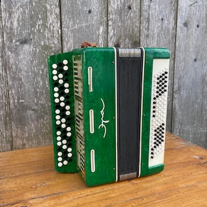 May include: A green accordion with white and black accents. The instrument has a rectangular shape with multiple buttons and a bellows design. The accordion is set against a wooden background.