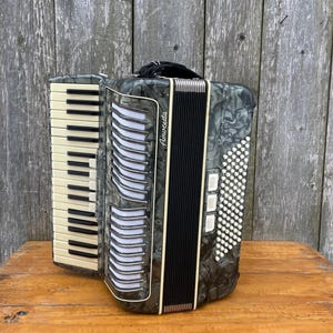 May include: A vintage Amoretta accordion with a black and gray marbled finish. The accordion features ivory keys, black and white buttons, and a black handle. The instrument is set against a weathered wooden background.