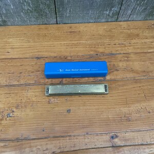 May include: A brass harmonica and a blue case are displayed on a wooden surface. The case is labeled "Swan Musical Instrument." The harmonica is rectangular with a brass finish. The background is a weathered wooden wall.