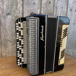 May include: A black accordion with white and cream-colored accents. The instrument features a keyboard with white buttons and a bellows with black pleats. The name "Кировский" is written in cursive on the front. The accordion is set against a wooden background.