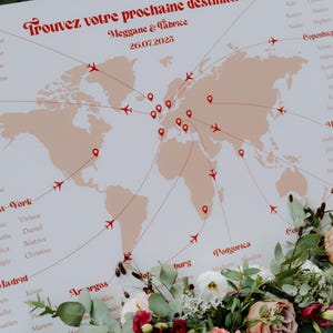 Op de afbeelding: Een trouwkaart met een wereldkaartontwerp, met rode vliegtuigpictogrammen en bestemmingslijnen. De tekst luidt "Trouvez votre prochaine destination" en "Meggane & Fabrice 26.07.2025". Versierd met een bloemstuk.
