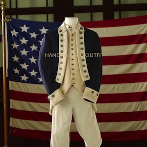 May include: A navy blue and cream-colored historical outfit, including a jacket, vest, and pants, displayed in front of an American flag. The jacket has cream-colored cuffs and lapels with decorative buttons. The text "HANDMADE OUTFIT" is visible on the jacket.