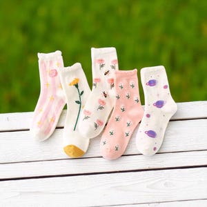 May include: Five pairs of colorful socks with various designs. One pair has pink stripes and a star, another has a yellow flower, and a third has pink flowers and a bee. The fourth pair is pink with small white flowers, and the last pair has a planet design.
