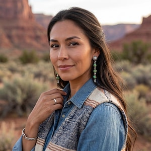 May include: A woman wearing long dangle earrings with turquoise beads and silver accents. The earrings are paired with a silver bracelet and ring. She is wearing a denim shirt and a patterned vest.