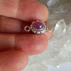 May include: A silver jewelry clasp featuring a central oval cabochon-cut deep pink gemstone. The metal setting has a scalloped edge design with a small circular loop on one side and a flat rectangular tab on the other for fastening.