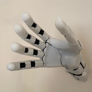 May include: A light gray robotic hand with articulated fingers. The fingers have black joints and white tips. The hand is posed in an open position, with the palm facing the viewer. The hand is made of plastic.
