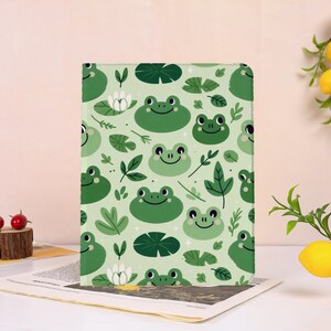 Op de afbeelding: Een groen notitieboek met een herhaald patroon van lachende kikkers, waterlelies en bladeren. De achtergrond is lichtgroen. Het notitieboek staat op een bedrukte pagina.