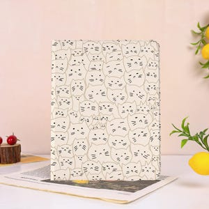 May include: A rectangular notebook with a cream-coloured cover featuring a repeating pattern of black line-drawn cat faces. The cats have various expressions, including closed eyes, straight mouths, and whiskers, covering the entire front surface.