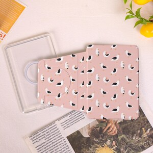 May include: A pink tablet case with a pattern of white and black seagulls. The case is open, revealing a clear plastic holder and a patterned cover. The design is repeated on the cover, which is folded open.