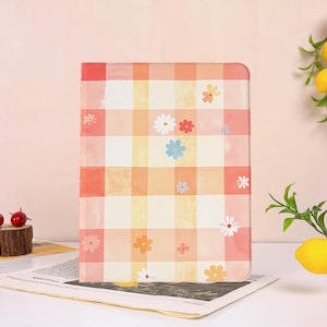 May include: A rectangular notebook with a colorful plaid pattern in shades of orange, yellow, and white. The notebook is decorated with various floral designs. The notebook is sitting on top of a newspaper.