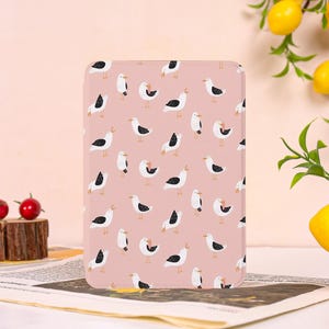 May include: A rectangular, pink tablet case featuring a repeating pattern of cartoon seagulls in black, white, and yellow. The seagulls are in various poses, with some appearing to be squawking. The case is on a newspaper.