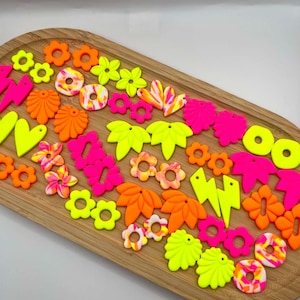 May include: A collection of assorted neon-coloured polymer clay earring components in shapes including flowers, hearts, lightning bolts, and leaves. The pieces feature solid bright pink, orange, and yellow hues alongside marbled patterns of these colours.
