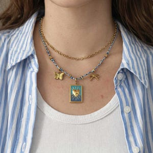 Peut inclure: Un collier superposé avec des chaînes et des pendentifs dorés. Le collier inférieur a un cordon bleu et blanc avec un papillon, une libellule et un pendentif rectangulaire avec un motif de cœur. La personne porte un débardeur blanc et une chemise rayée bleue et blanche.