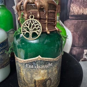 以下が含まれることがあります： 「Ent-draught」と書かれた装飾的なラベルと木のペンダントが付いた緑色のガラス瓶。ボトルは茶色のアクセントと金色のワックスシールで飾られています。ラベルには素朴な森の風景が描かれています。