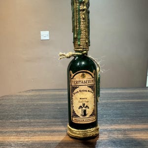 Könnte beinhalten: Eine grüne Glasflasche mit einem goldenen und grünen Wachssiegel. Die Flasche ist mit "Veritaserum Truth Telling Serum" beschriftet und hat ein Vintage-Design. Die Flasche ist mit Schnur gebunden und trägt ein Premium-Qualitätslabel.