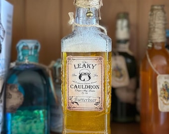 Könnte beinhalten: Eine Glasflasche mit goldener Flüssigkeit und einem Etikett mit der Aufschrift "The Leaky Cauldron Butterbeer". Die Flasche ist mit Schnur umwickelt und hat eine kleine Glocke, die oben hängt.