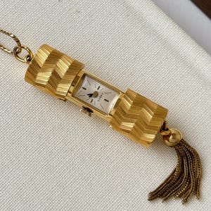 May include: A gold-tone pendant watch featuring a rectangular white face with black hands and the text "FRECO". The watch case is flanked by two textured gold-tone cylindrical components with a chevron pattern and a gold-tone chain tassel at the base.