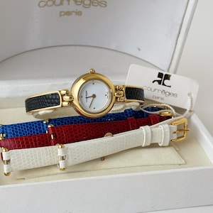 May include: A gold Courrèges Paris watch with a white face and black leather strap, displayed in a white box. Includes spare watch straps in red, blue, and white.