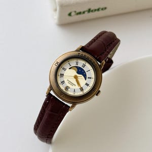 May include: A wristwatch with a dark brown embossed leather strap and a round, antique-gold tone metal case. The dial features Roman numerals, a moon phase display with stars, gold hands, and the text TORNADO QUARTZ with a date window at six.