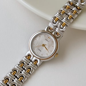 Puede incluir: Un reloj Seiko Delfina con una pulsera plateada y dorada. La esfera del reloj es de color claro con agujas e indicadores dorados. La correa del reloj presenta un patrón repetitivo de eslabones plateados y dorados.