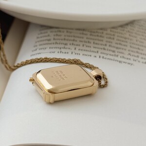 Puede incluir: Un collar con colgante rectangular dorado con una cadena delicada. El colgante tiene un dise&ntilde;o con bisagras y parece estar grabado con texto. El collar descansa sobre las p&aacute;ginas abiertas de un libro.